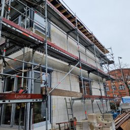 P&B Consulting Paweł Boniek - Budowa domu jednorodzinnego z widocznymi oknami i roletami zewnętrznymi w trakcie montażu, otoczona rusztowaniem Layher, z widocznym budynkiem z czerwonej cegły w tle i samochodem dostawczym z boku.