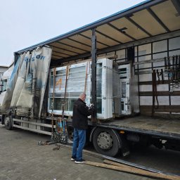 P&B Consulting Paweł Boniek - Mężczyzna w czarnej kurtce i jeansach rozładowuje z ciężarówki białe ramy okienne zapakowane w folię stretch; widoczne pasy mocujące i narzędzia zabezpieczające ładunek.