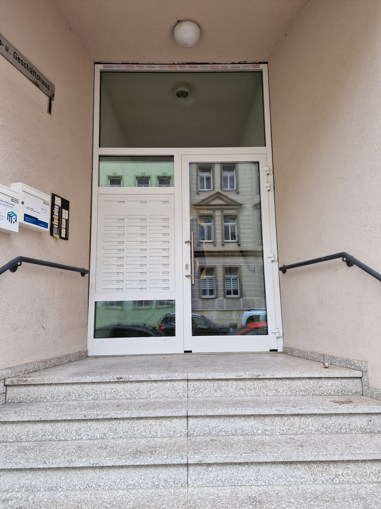 Białe drzwi wejściowe do kamienicy w Lipsku, Niemcy. Drzwi z przeszkleniami i skrzynkami pocztowymi, widoczne odbicie budynku w szybach. Zamontowane przy schodach wejściowych.