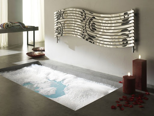 Minimalistyczna łazienka z wpuszczoną w podłogę wanną wypełnioną pianą, dekoracyjnym grzejnikiem drabinkowym z abstrakcyjnym wzorem na ścianie, płonącymi świecami i rozsypanymi płatkami róż.