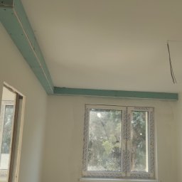 SELMI UB ANDRII SOLOVIOV - Wnętrze w trakcie prac: widoczne płyty kartonowo-gipsowe zamontowane na suficie, okno z widokiem na zewnątrz i stos płytek gresowych na podłodze.