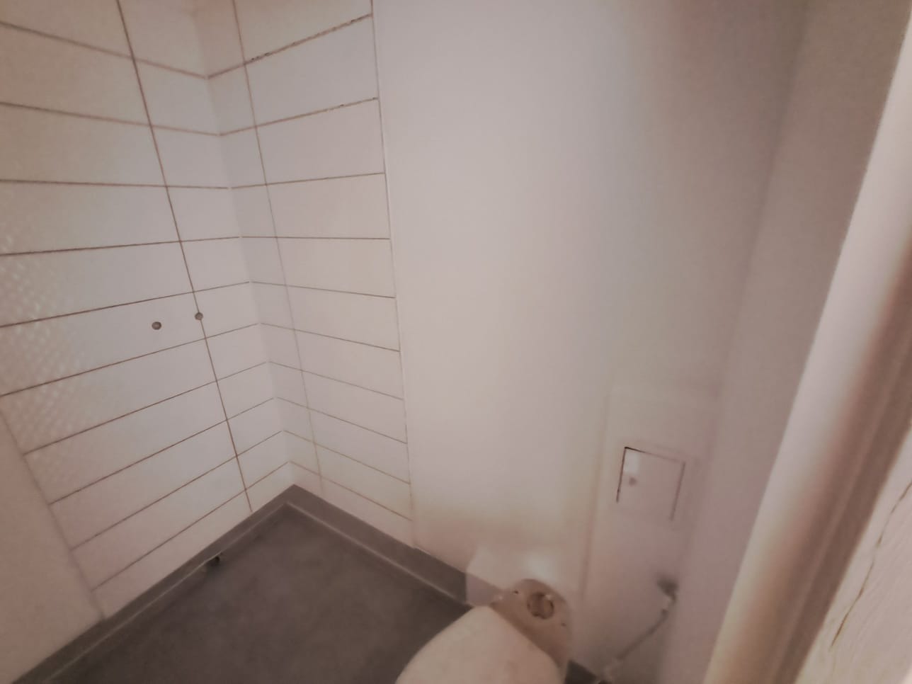 Wnętrze łazienki w trakcie prac wykończeniowych, widoczne ułożone białe płytki na ścianach, szara podłoga, częściowo zamontowana toaleta i biała ściana z dostępem rewizyjnym.