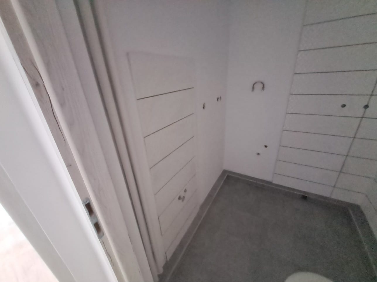 Wnętrze łazienki w trakcie prac wykończeniowych: widoczna zabudowa z drzwiczkami, częściowo ułożone płytki na ścianie z widocznymi otworami na instalacje, szara posadzka.