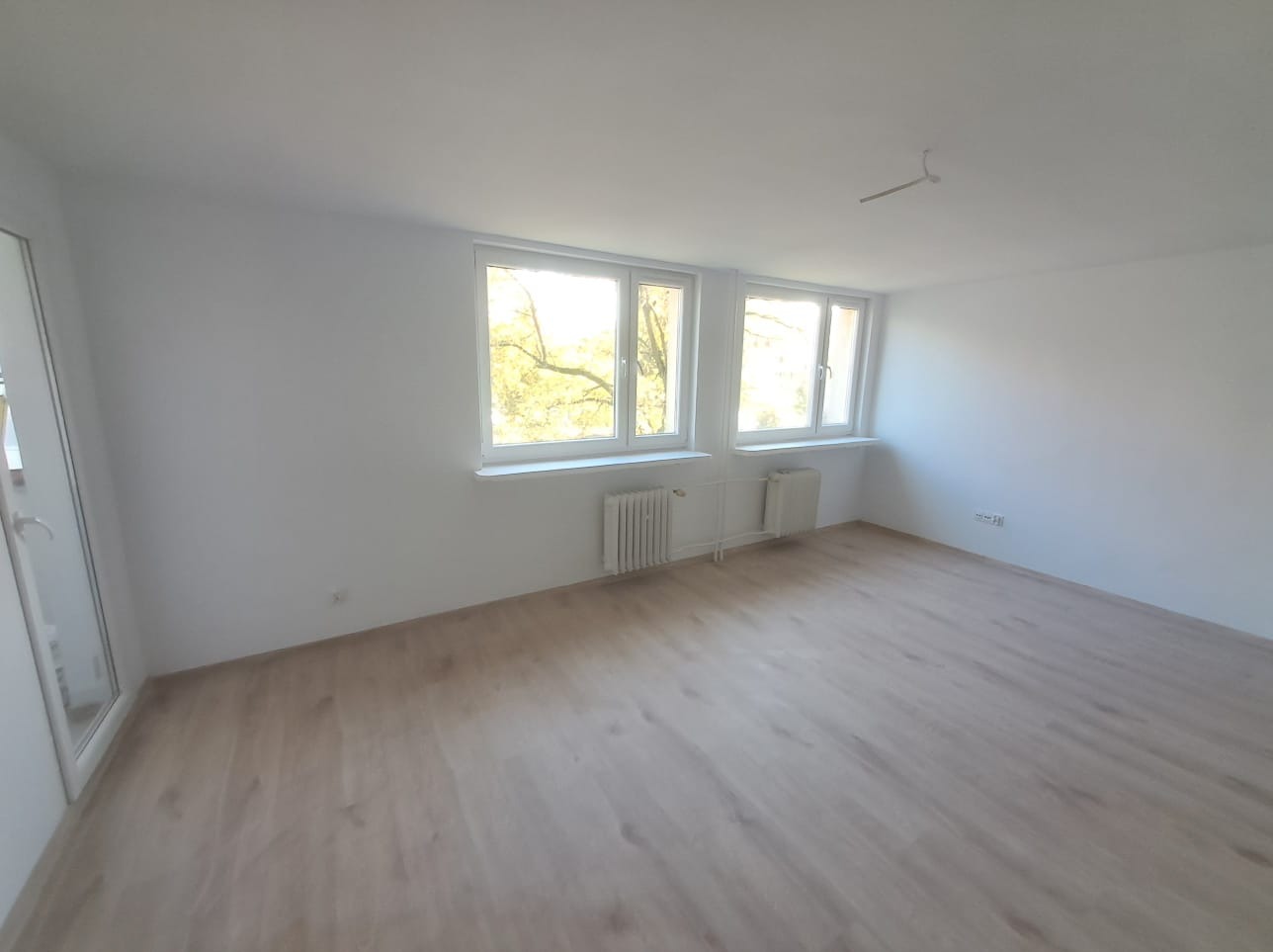 Pusty, jasny pokój po remoncie z panelami podłogowymi, białymi ścianami, dwoma oknami z widokiem na drzewa i grzejnikami pod oknami.