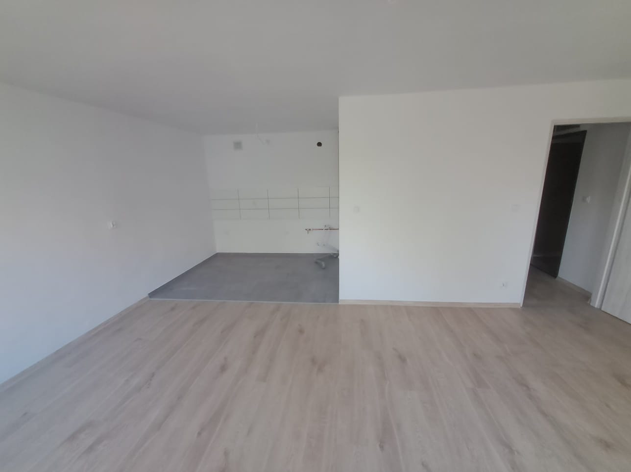 Pusty pokój w trakcie wykańczania: świeżo położone jasne panele podłogowe, surowy beton na podłodze w miejscu przyszłej kuchni, białe ściany przygotowane do malowania, widoczne punkty instalacji.