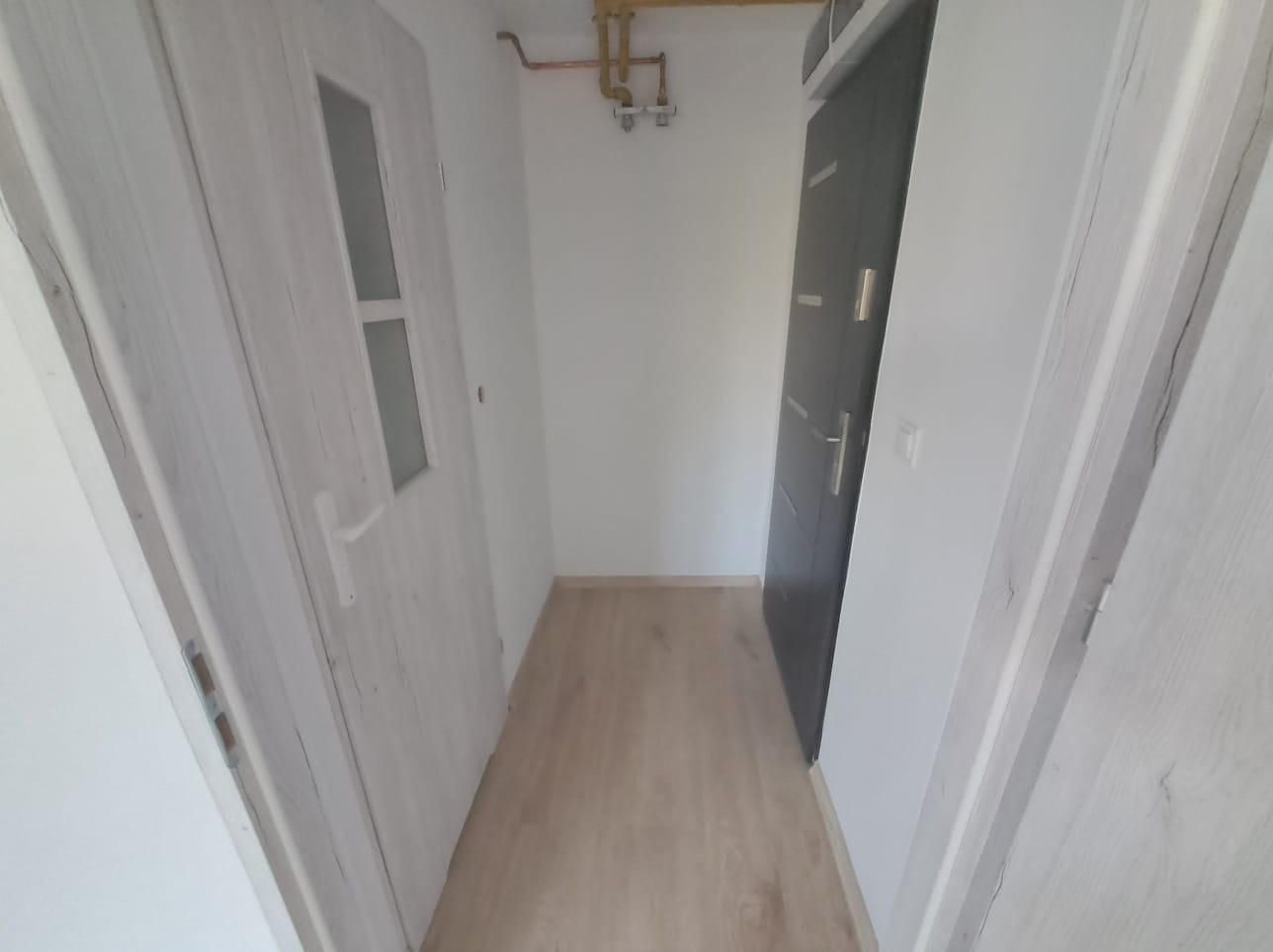 Korytarz po remoncie z panelami podłogowymi w odcieniach beżu, widoczne dwie ościeżnice z drzwiami: biała z przeszkleniem i grafitowa z poziomymi zdobieniami, na ścianie widoczna instalacja gazowa.