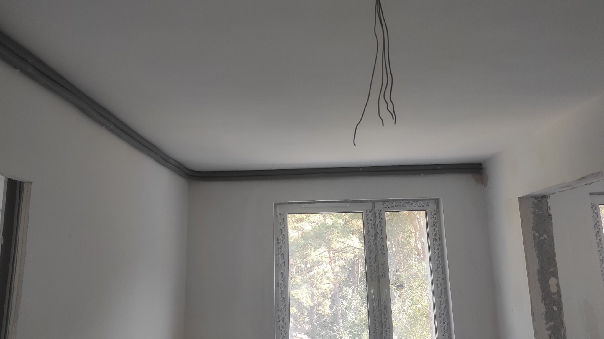 Surowe wnętrze w trakcie prac: szare rury prowadzone pod sufitem wzdłuż ściany, zwisające kable elektryczne, okno z widokiem na las, ściany przygotowane pod malowanie.