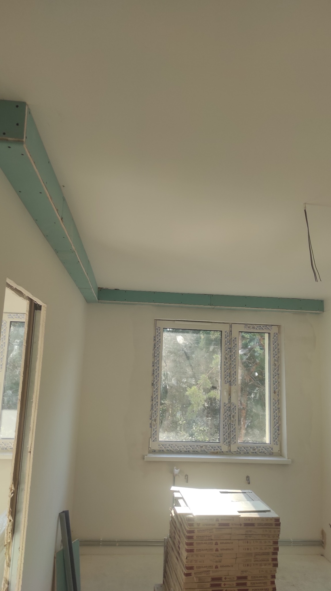 Wnętrze w trakcie prac: widoczne płyty kartonowo-gipsowe zamontowane na suficie, okno z widokiem na zewnątrz i stos płytek gresowych na podłodze.