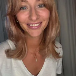 Portret uśmiechniętej kobiety o blond włosach z grzywką, złotych kolczykach i naszyjnikiem z zawieszką w kształcie litery 'E', w białym t-shircie na tle zasłony.