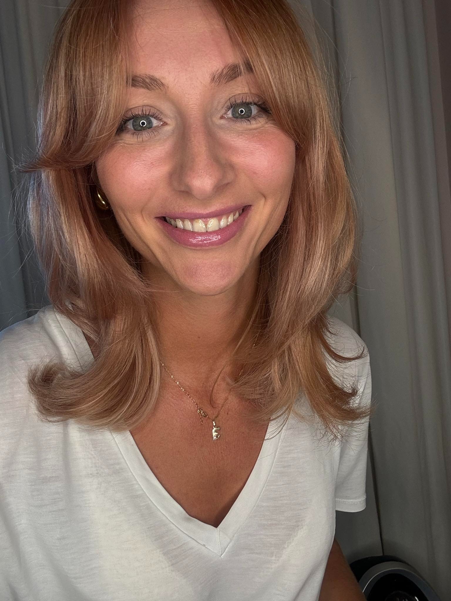 Portret uśmiechniętej kobiety o blond włosach z grzywką, złotych kolczykach i naszyjnikiem z zawieszką w kształcie litery 'E', w białym t-shircie na tle zasłony.