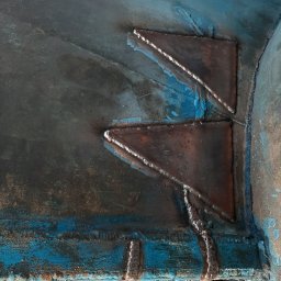 Emilia Stochaj - Dwa trójkątne wsporniki przyspawane do niebieskiej, pomalowanej powierzchni, widoczne ślady spawania i odpryski farby, maska spawalnicza w tle.