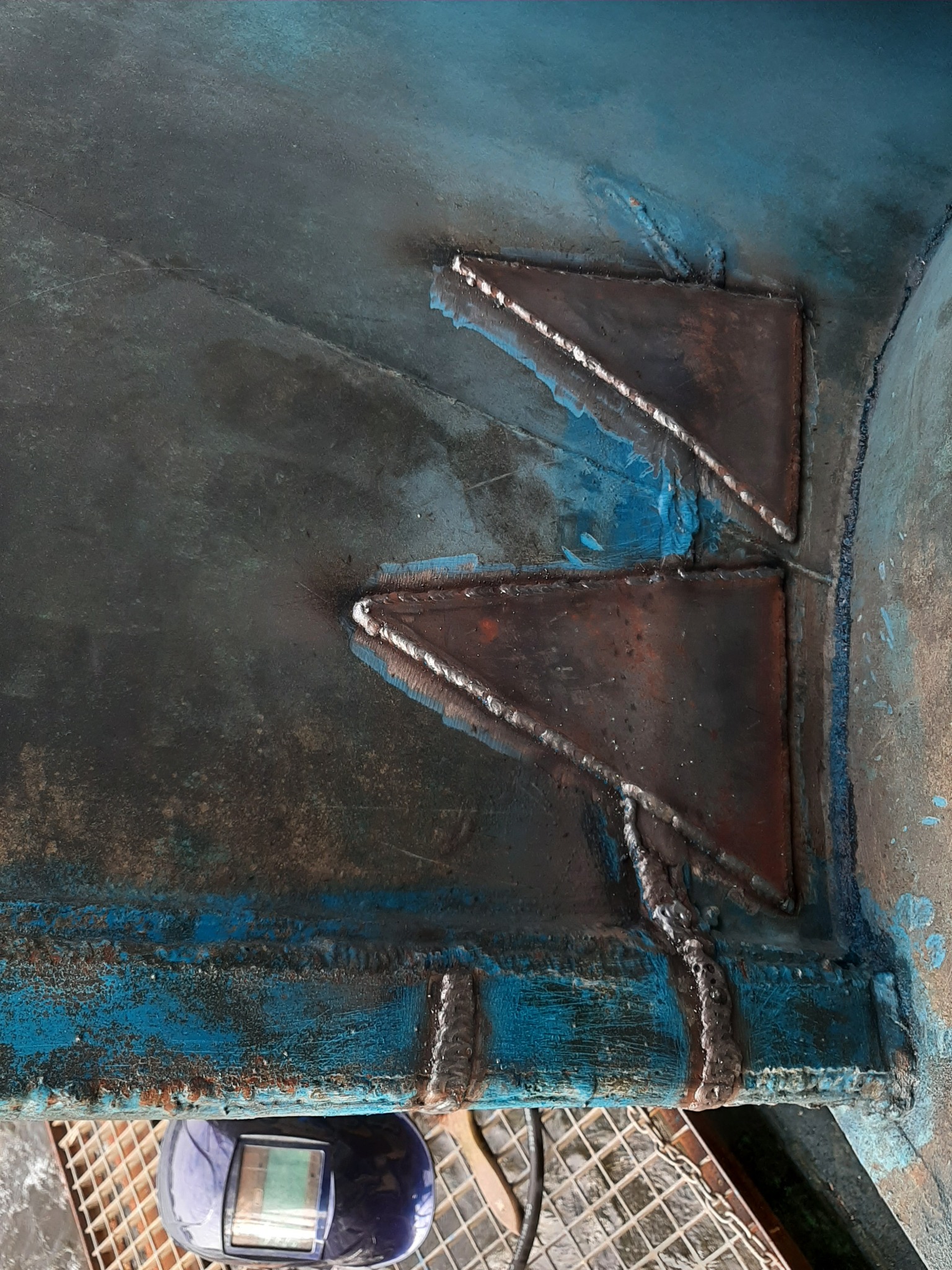 Dwa trójkątne wsporniki przyspawane do niebieskiej, pomalowanej powierzchni, widoczne ślady spawania i odpryski farby, maska spawalnicza w tle.