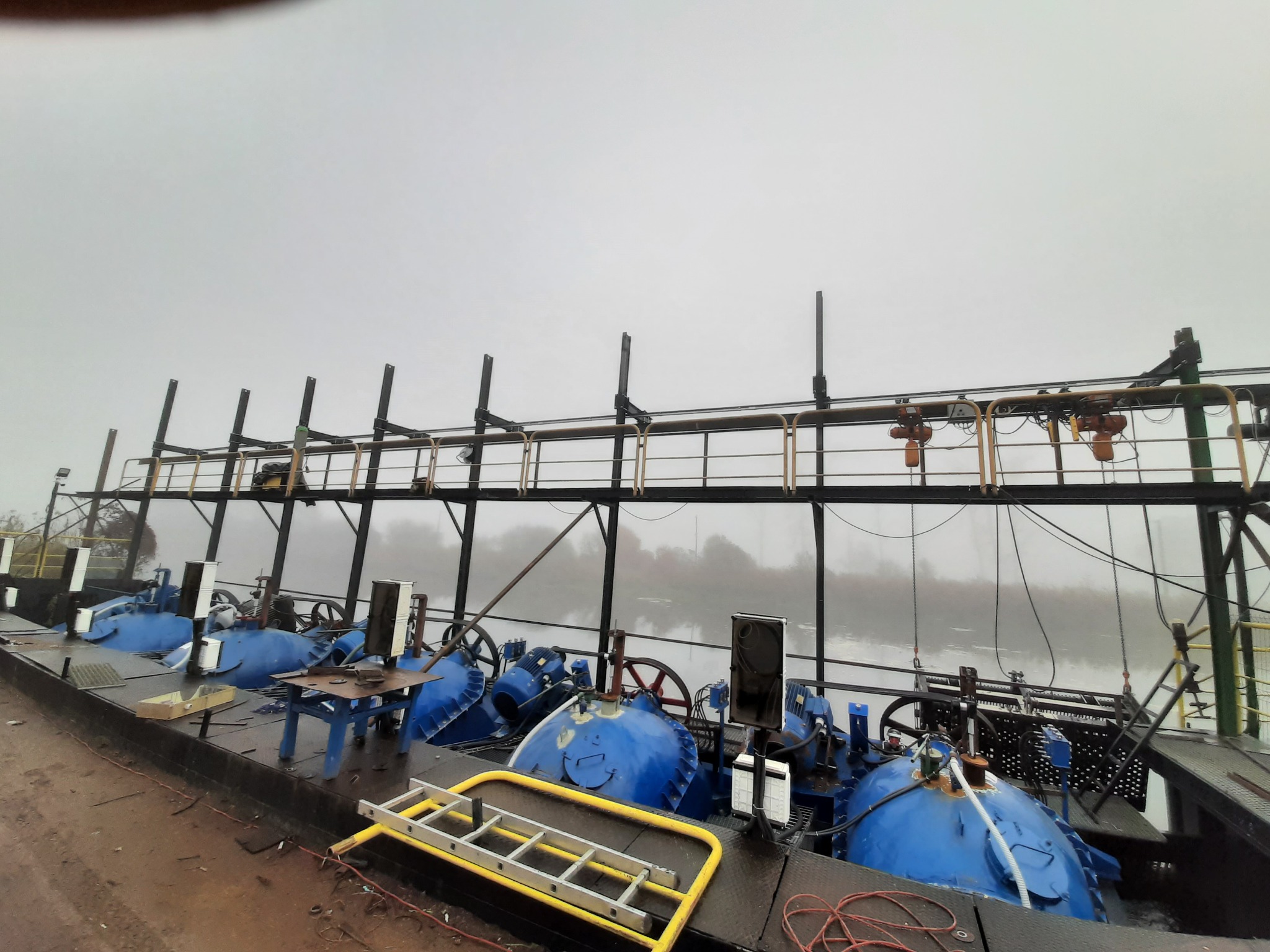 Zestaw niebieskich pomp przemysłowych z silnikami, umieszczony na platformie z metalową konstrukcją w tle zamglonego krajobrazu.