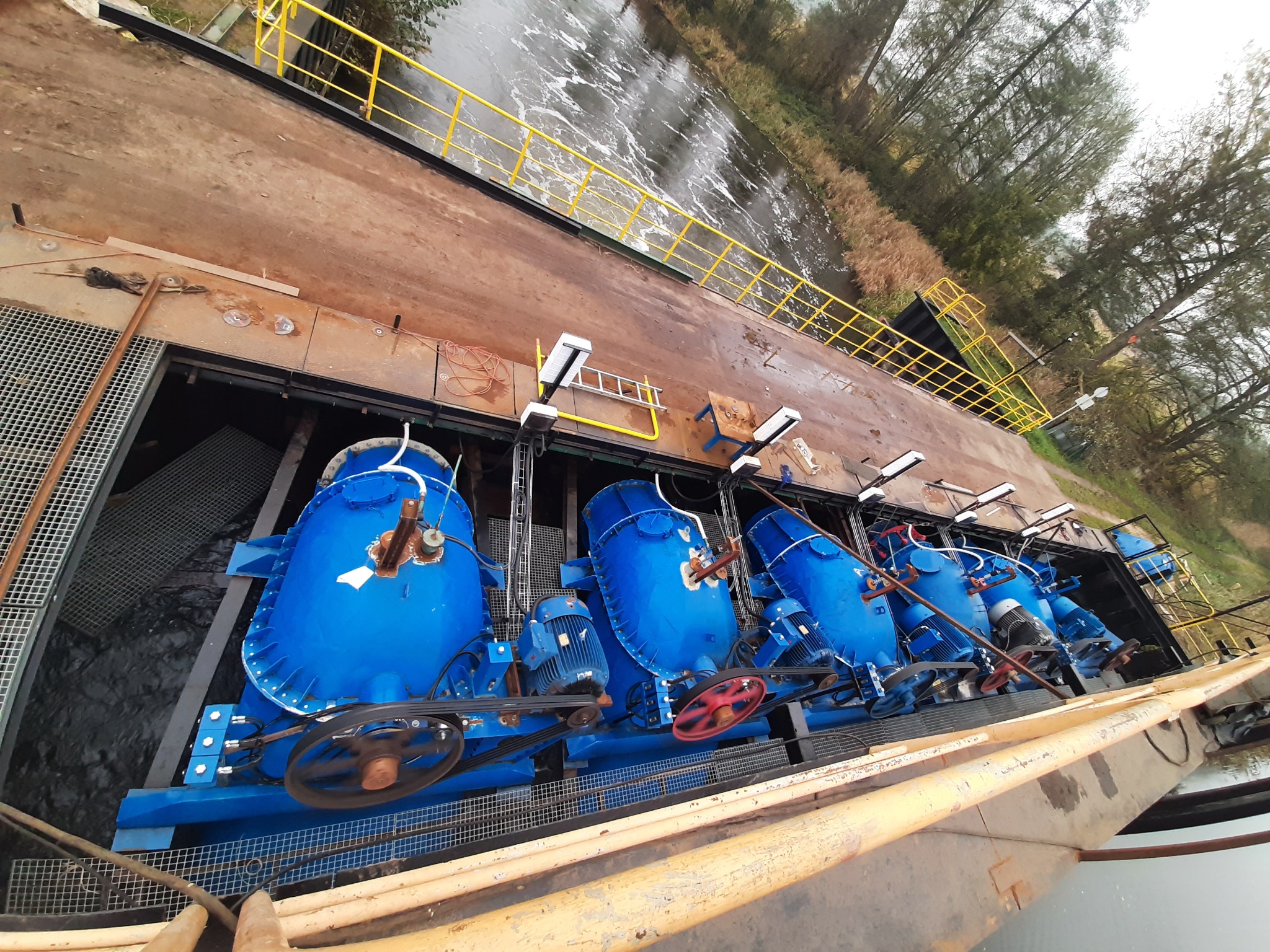 Kompleksowe urządzenia w niebieskiej obudowie z silnikami elektrycznymi i przekładniami pasowymi, umieszczone na metalowej platformie nad zbiornikiem wodnym, otoczone barierkami ochronnymi.