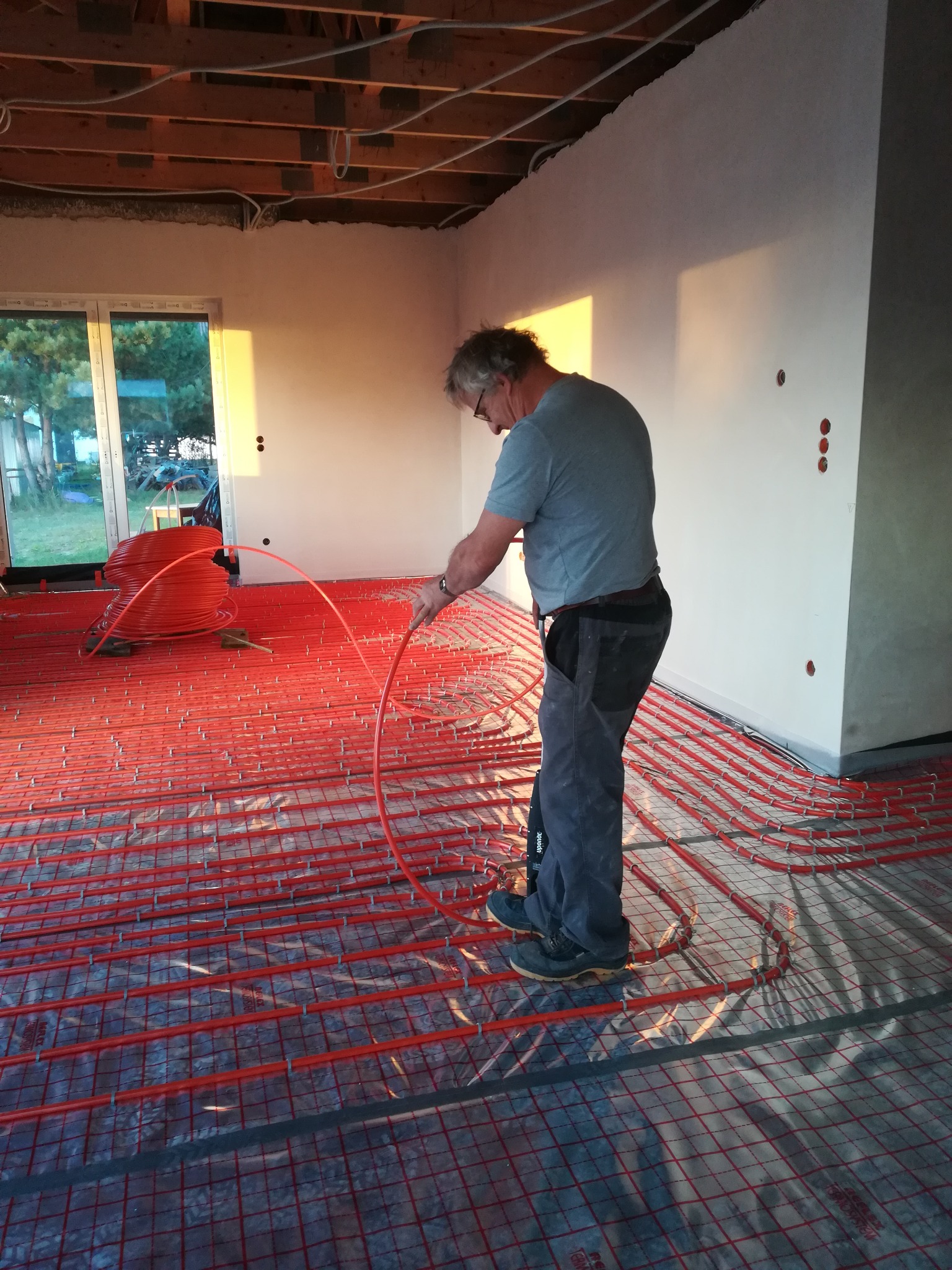 Instalacja ogrzewania podłogowego: mężczyzna układa czerwone rury grzewcze na folii z siatką w surowym stanie, widoczne okno z zachodzącym słońcem.
