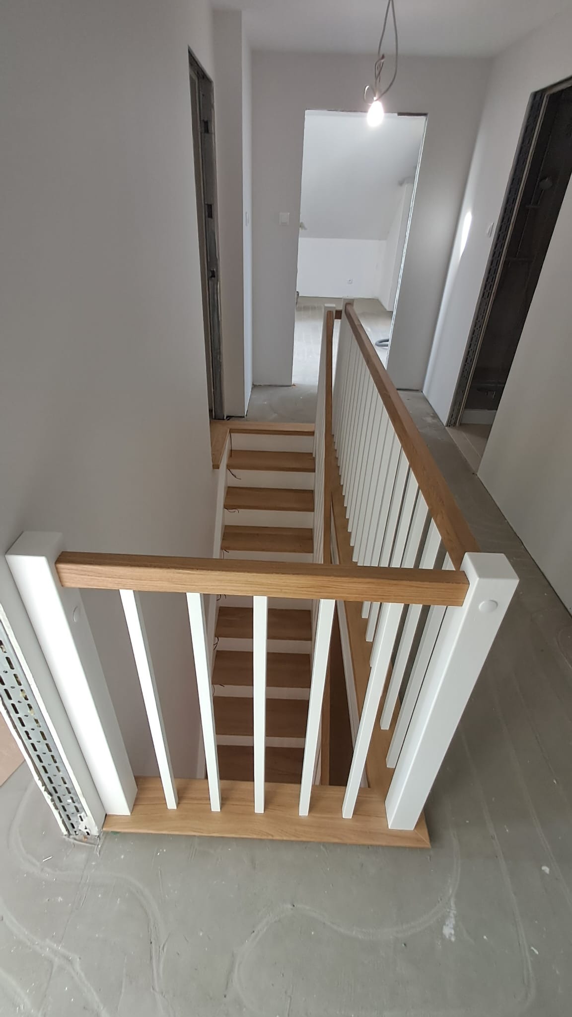 Jasne, drewniane stopnie schodów z białymi balustradami w surowym wnętrzu. Widok z góry na klatkę schodową w trakcie wykończenia.