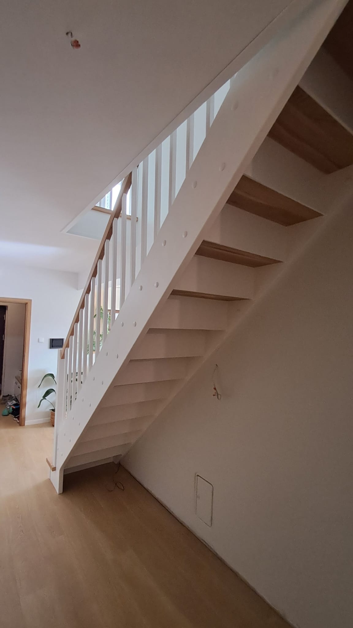 Białe schody z drewnianymi stopniami, widok z dołu, z fragmentem podłogi i ściany. Minimalistyczny design, jasne wnętrze, widoczne detale wykończenia.