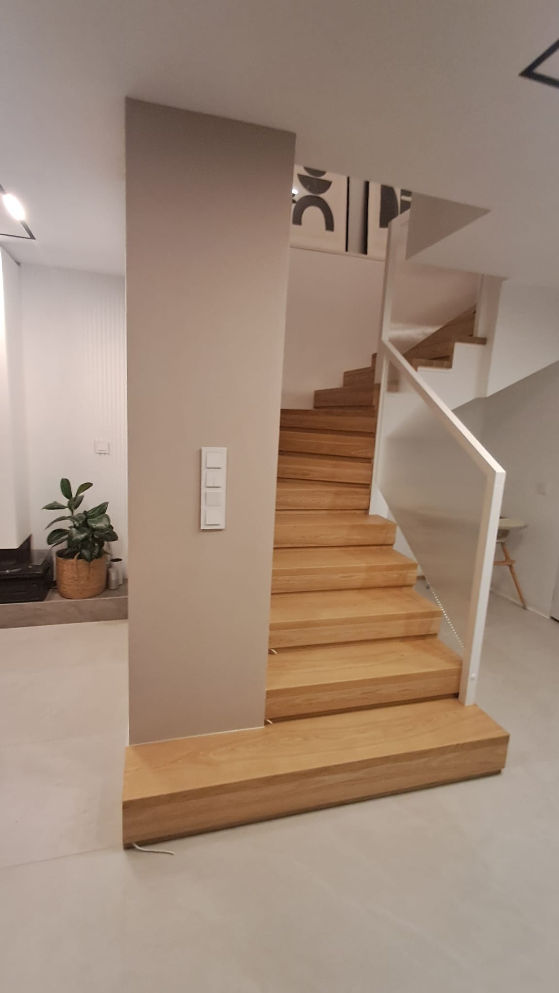 Jasne, drewniane stopnie schodów z białymi balustradami i szklanym wypełnieniem, w nowoczesnym wnętrzu z beżową kolumną i rośliną doniczkową w tle.