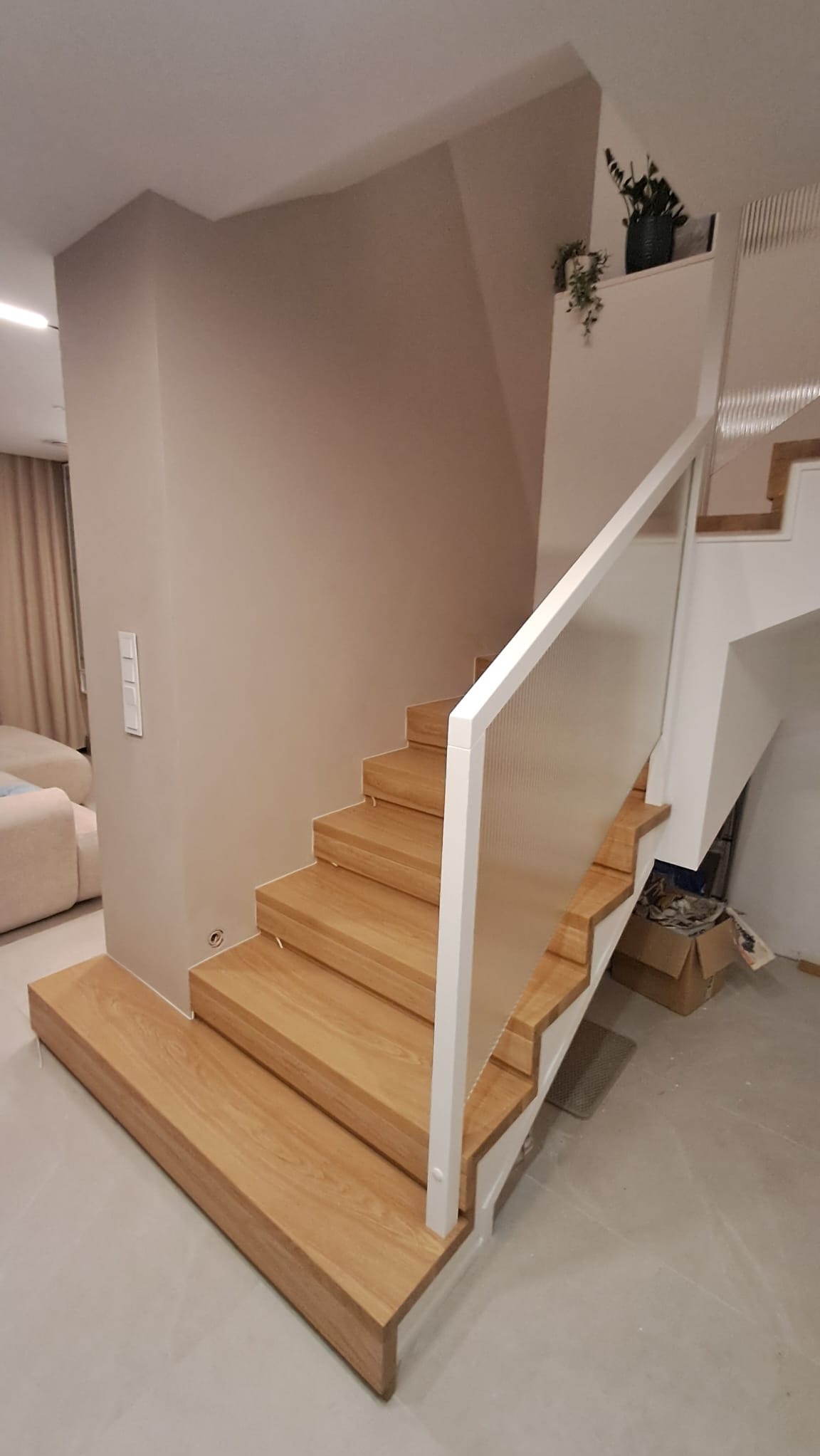 Jasne, drewniane stopnie z białymi balustradami i szklanym wypełnieniem, prowadzące do góry. Minimalistyczny design, neutralna kolorystyka ścian, widok z dołu.