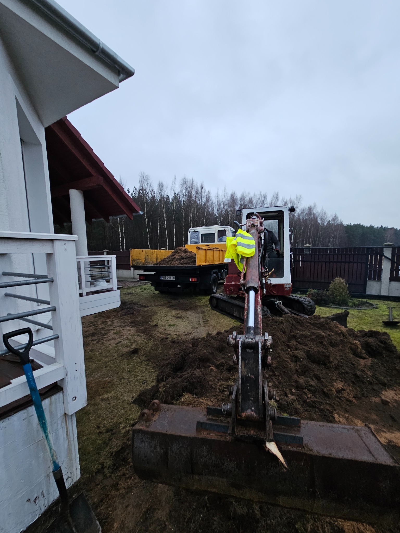Mini koparka z łyżką na pierwszym planie, w tle wywrotka z piaskiem, widok na teren budowy przy domu jednorodzinnym w pochmurny dzień.