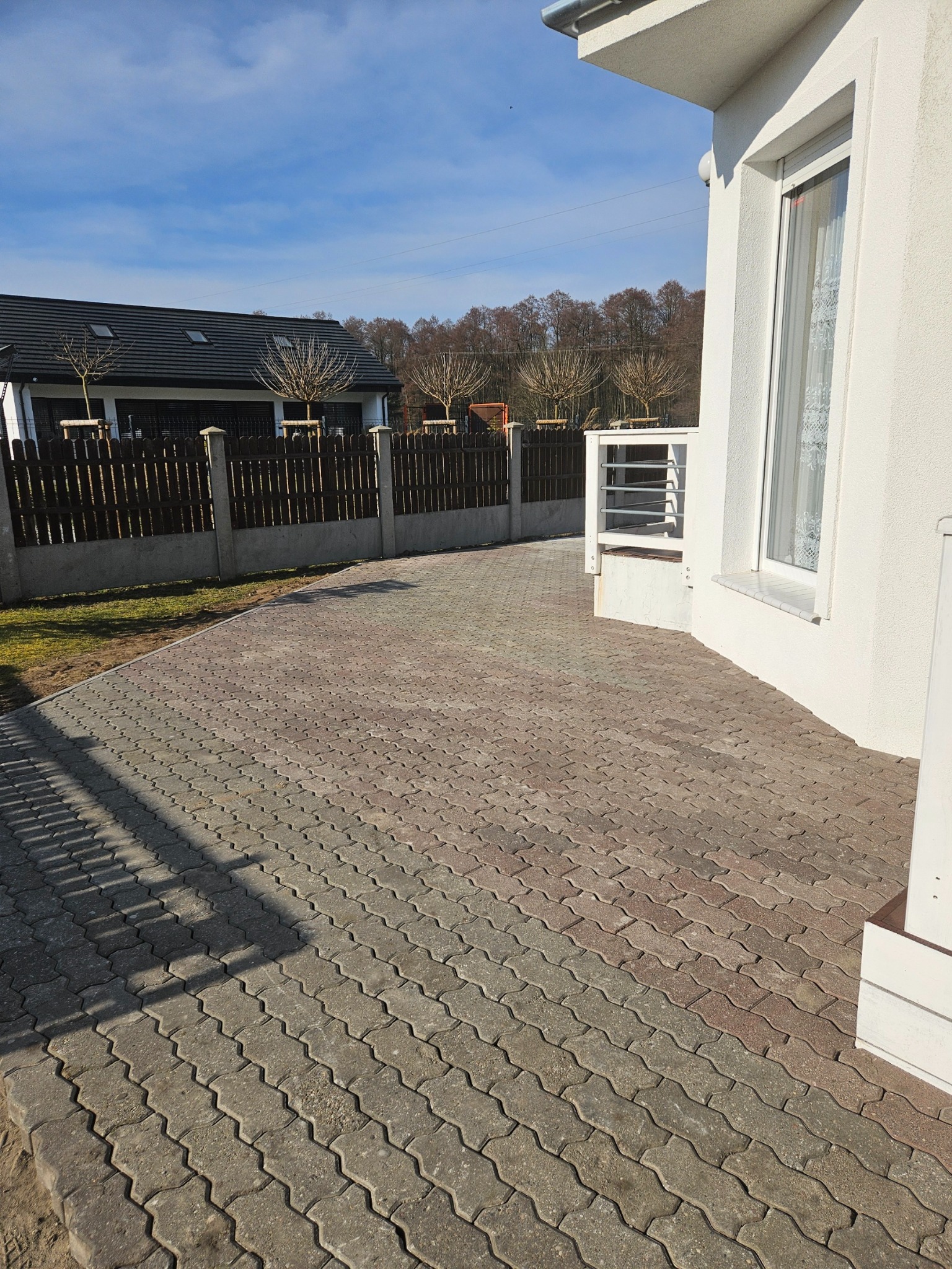 Ułożona szara kostka brukowa na podjeździe, widok na dom z białymi ścianami i drewniany płot w tle, słoneczny dzień.