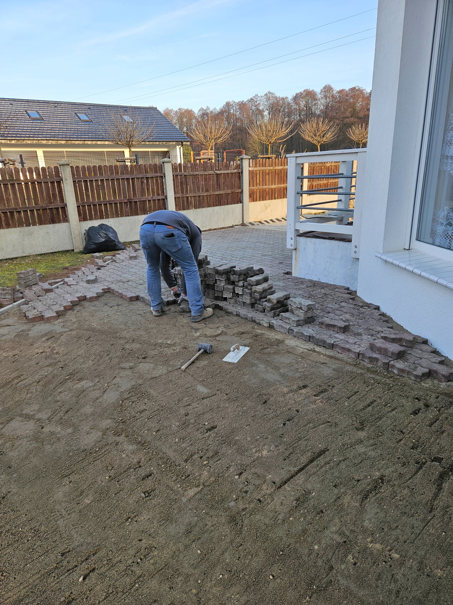 Pracownik w trakcie układania czerwono-szarej kostki brukowej na przygotowanym podłożu, widoczne narzędzia i częściowo ułożona nawierzchnia przed domem z drewnianym ogrodzeniem.