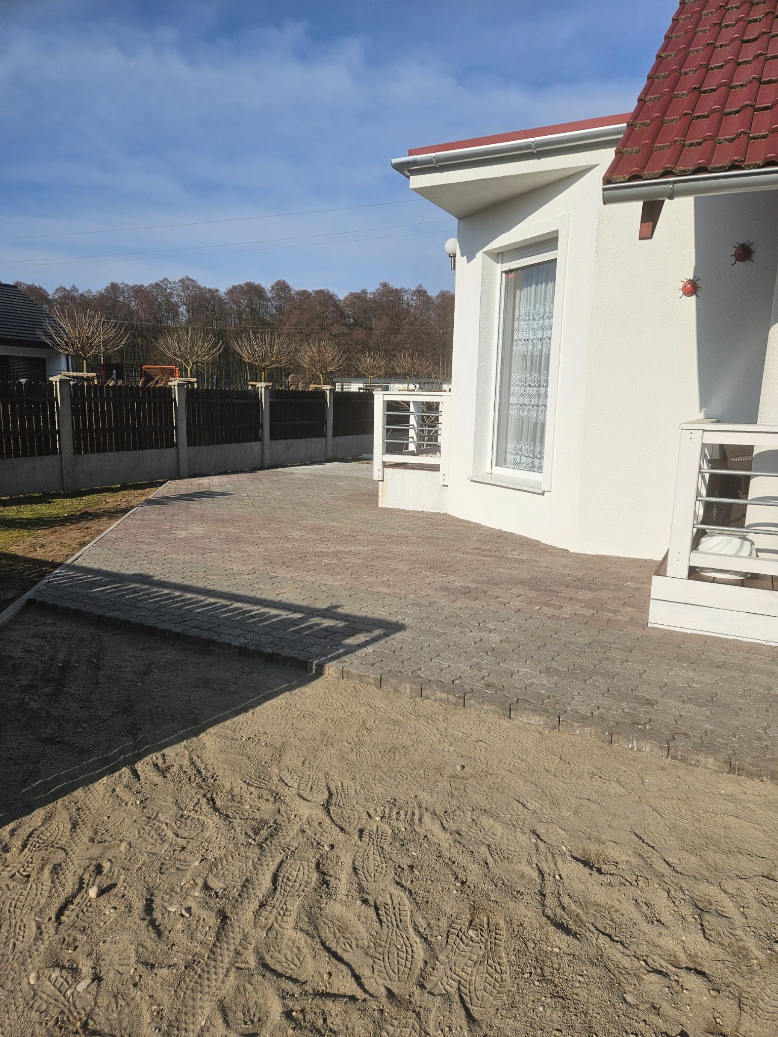 Ułożona kostka brukowa w odcieniach szarości i czerwieni przed białym domem z czerwonym dachem, widoczne ślady butów na piasku, ogrodzenie z drzewami w tle.