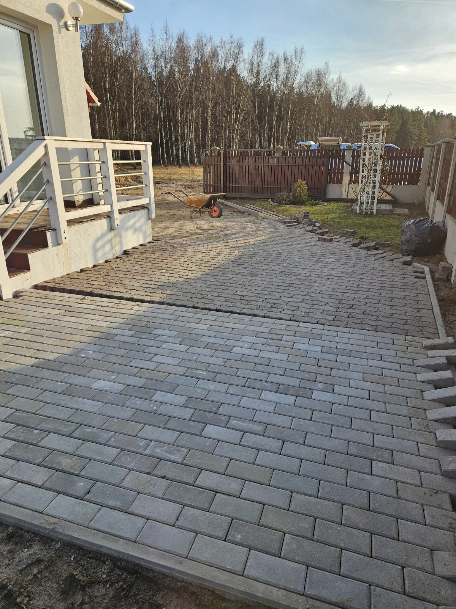 Układanie szarej kostki brukowej na podjeździe domu jednorodzinnego, widoczny etap prac z ułożoną częścią nawierzchni i przygotowanymi kostkami do dalszego układania, taczka w tle.