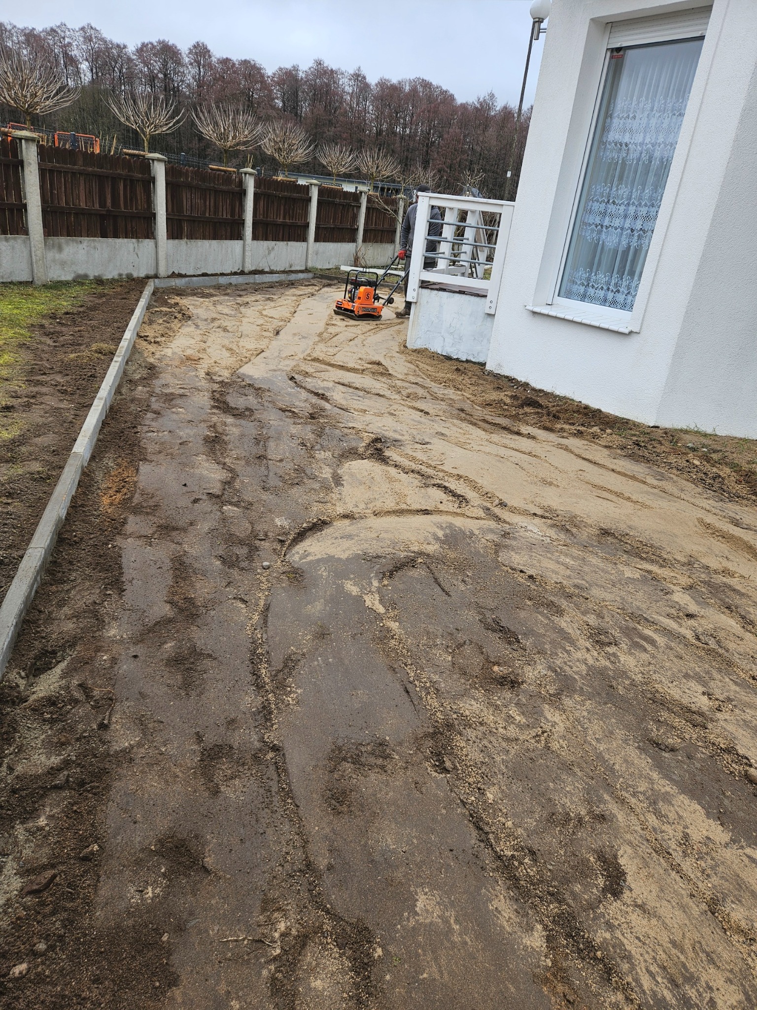 Przygotowanie podłoża pod nawierzchnię z kostki: zagęszczarka wibrująca ubija piasek na posesji z ogrodzeniem i fragmentem białego domu.
