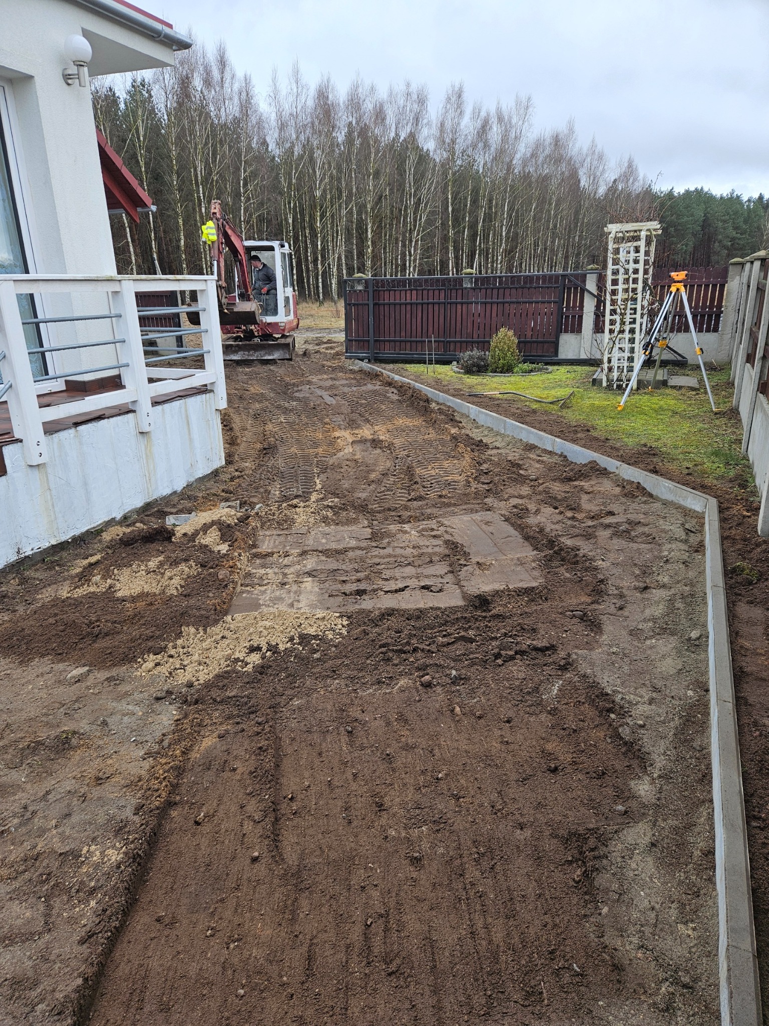 Przygotowanie terenu pod ścieżkę z obrzeżami, widoczny mini-koparka z operatorem, ziemia z wykopu i niwelator laserowy na statywie.