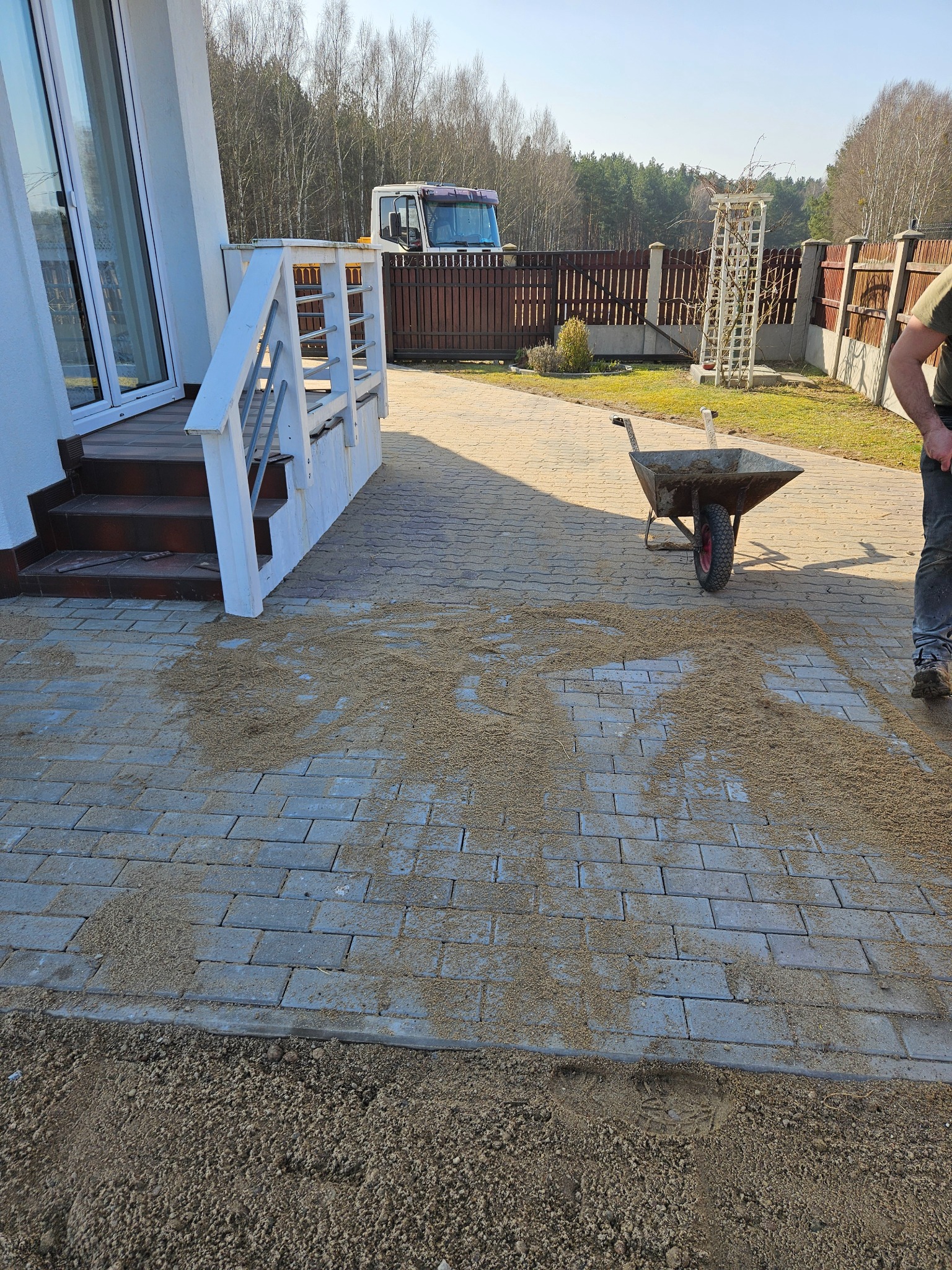 Szara kostka brukowa na podjeździe, częściowo zasypana piaskiem, z widoczną taczką i fragmentem sylwetki pracownika, w tle dom z białymi schodami i ciężarówka za ogrodzeniem.