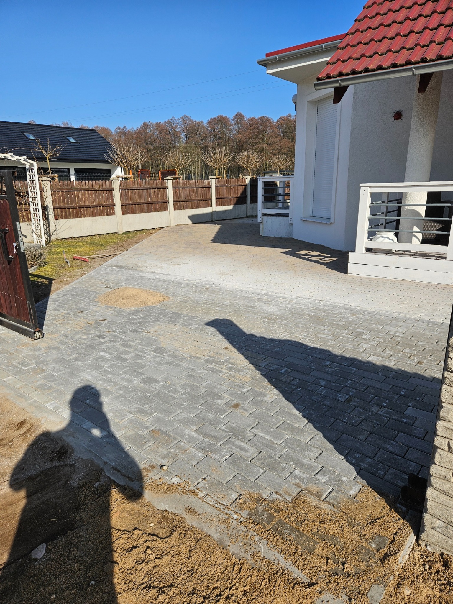 Układanie szarej kostki brukowej na podjeździe przed domem z czerwoną dachówką, widoczny cień osoby wykonującej prace i rozsypany piasek.