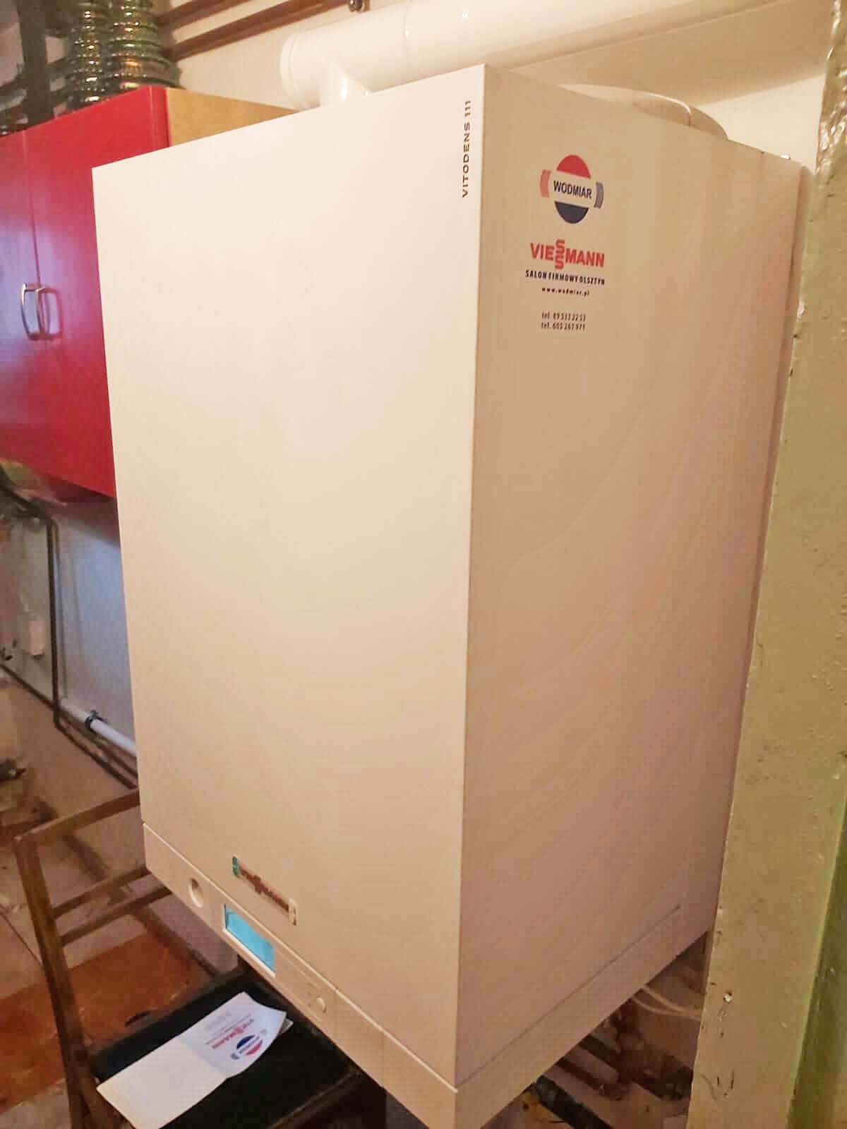 Biały piec gazowy Viessmann Vitodens 111 z logo Wodmar Salon Firmowy Olsztyn, zainstalowany w piwnicy z rurami i szafkami w tle.