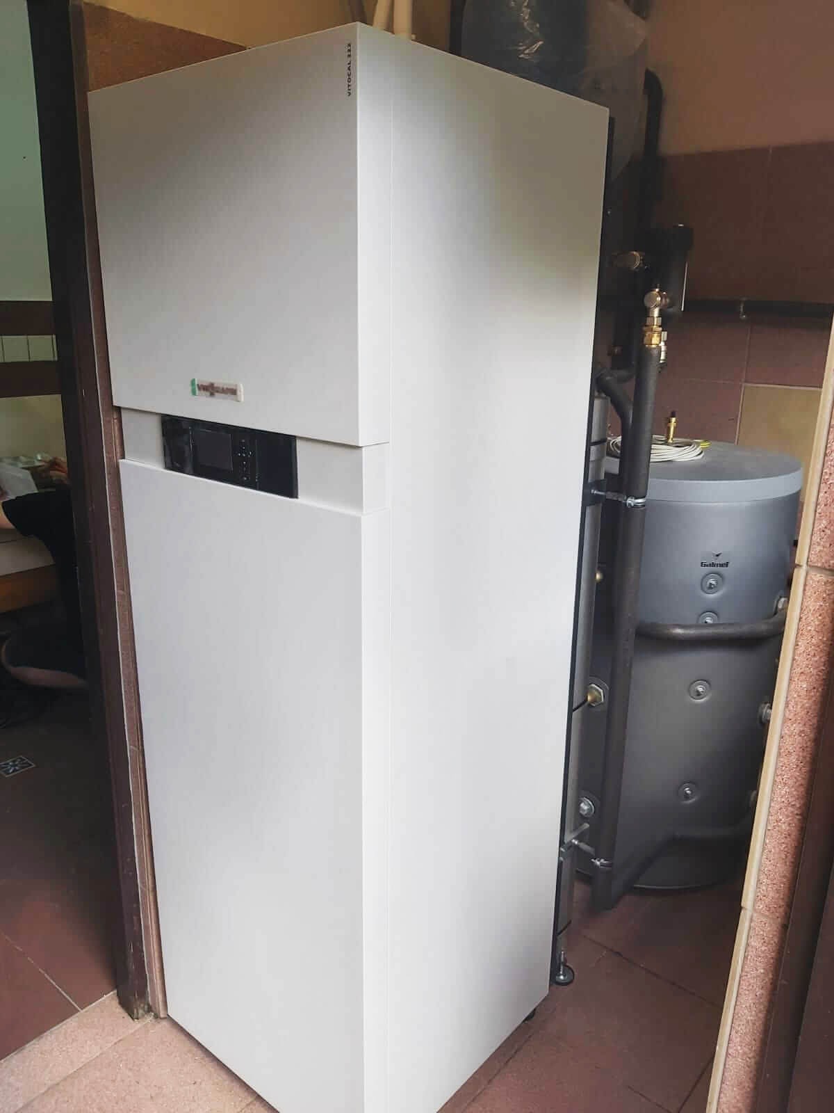 Biała, kompaktowa jednostka wewnętrzna z logo Viessmann, prawdopodobnie system grzewczy, z szarą obudową zasobnika ciepłej wody w tle, widoczne rury i instalacja.