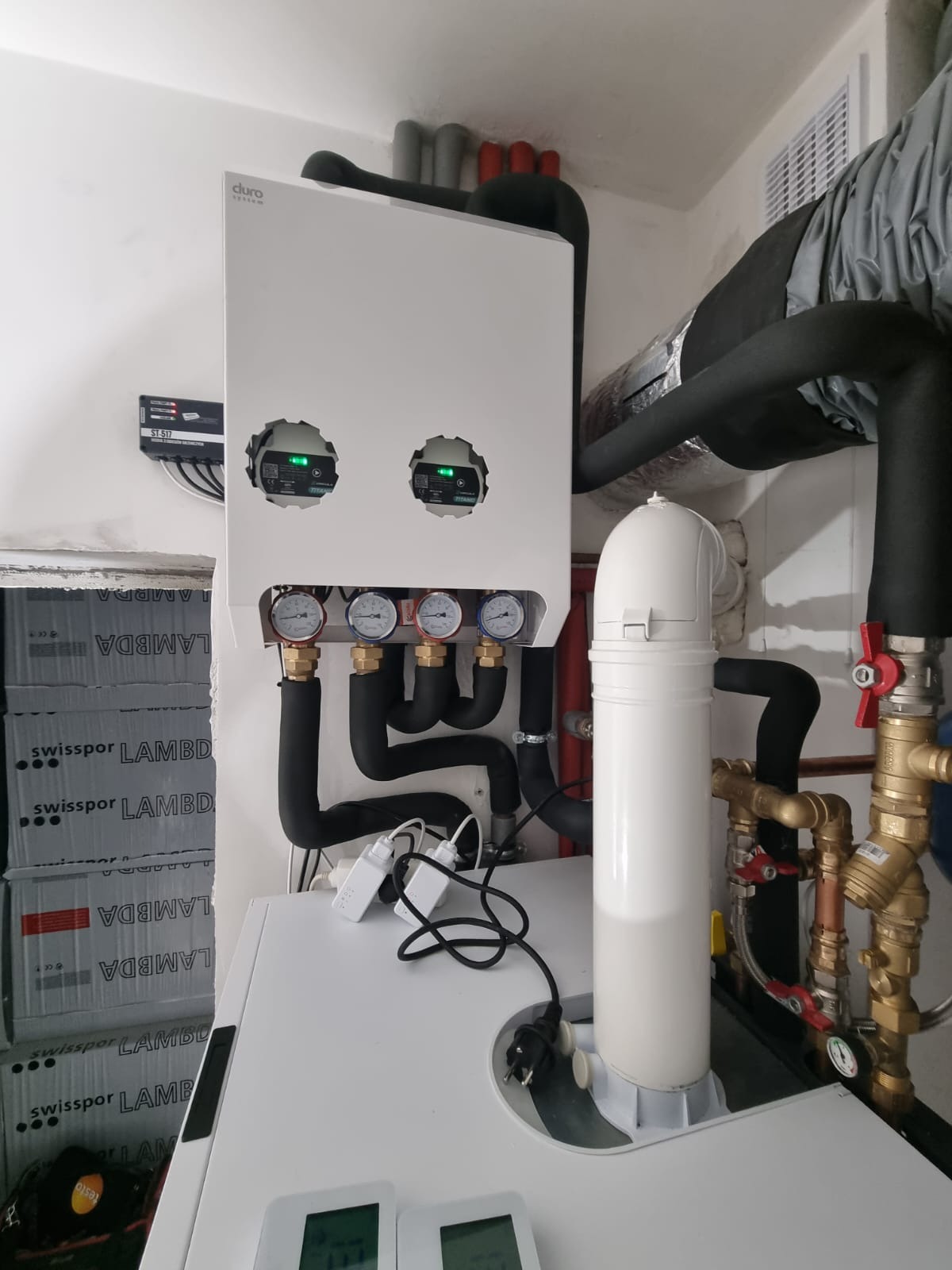 Nowoczesna, biała pompa ciepła marki Duro System z widocznymi manometrami i czarnymi rurami instalacyjnymi, zainstalowana w pomieszczeniu technicznym, obok widoczne opakowania izolacji Swissopor...