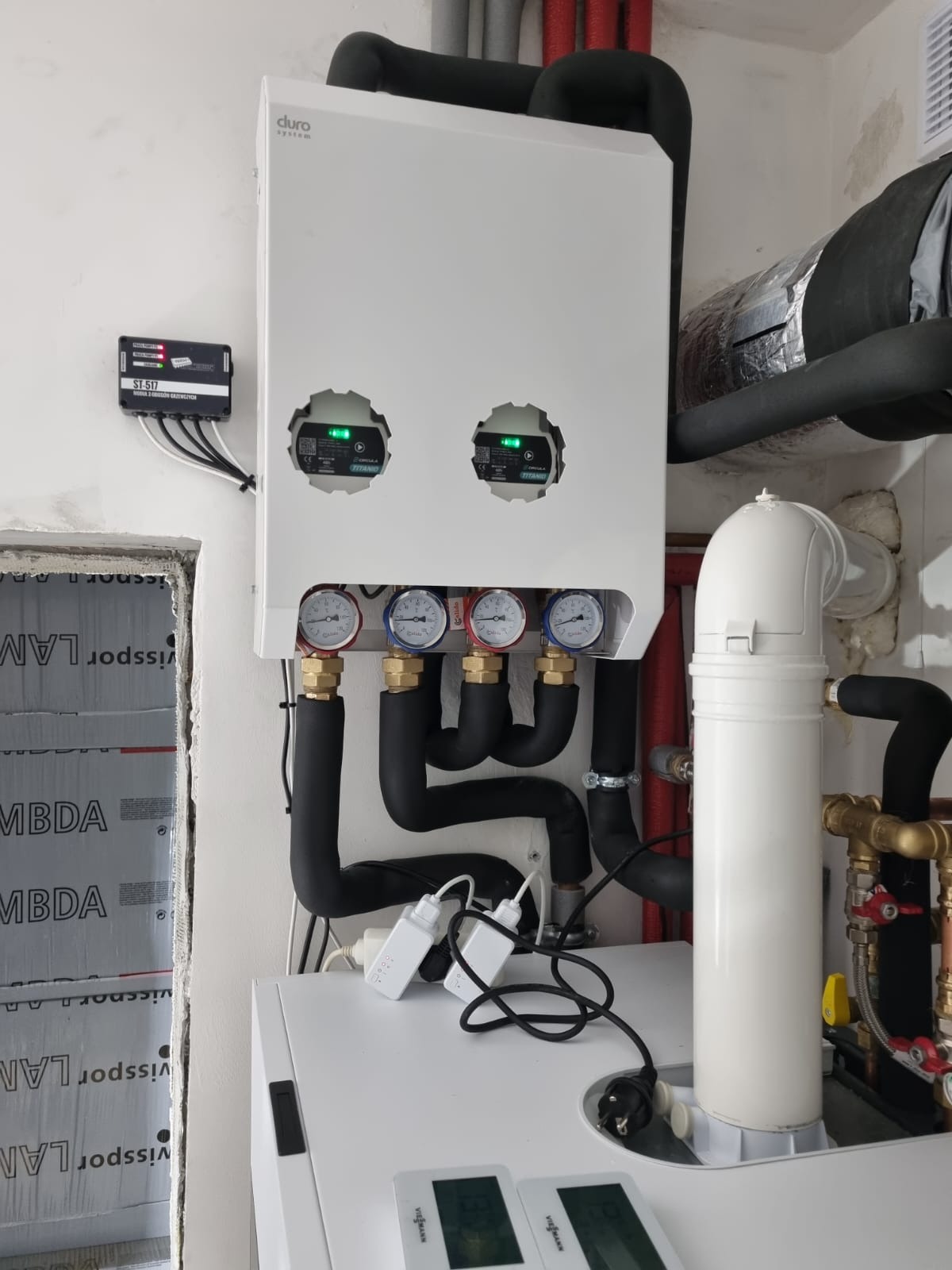 Biały piec grzewczy Duro System z dwoma okrągłymi wyświetlaczami i czterema manometrami, podłączony do czarnych rur, z dodatkowym sterownikiem ST-517 na ścianie i elementami instalacji Viessmann...