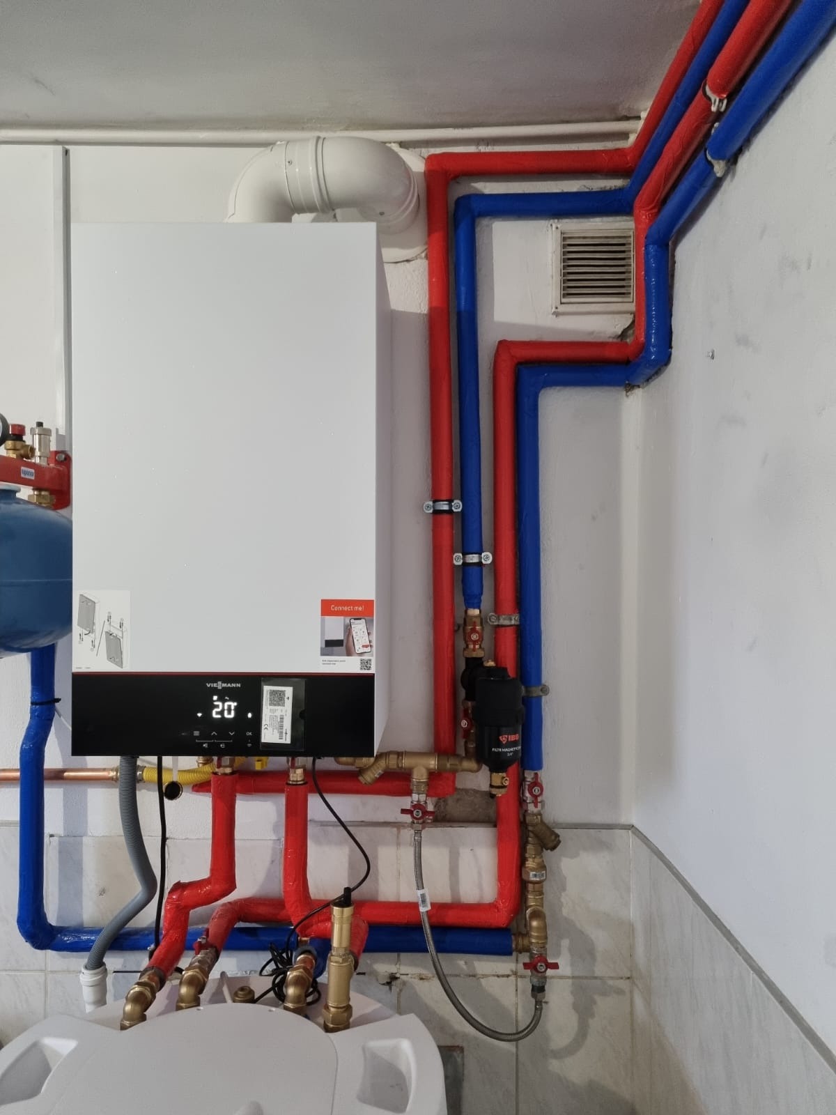 Nowoczesny, wiszący piec grzewczy Viessmann z podłączonymi rurami w kolorze czerwonym i niebieskim, zamontowany na białej ścianie z widoczną kratką wentylacyjną i elementami instalacji hydraulicznej.