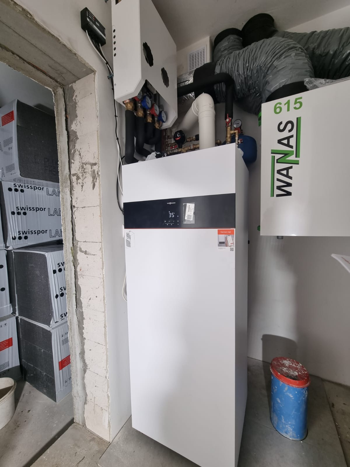 Biała instalacja grzewcza Viessmann z podłączonymi rurami i elementami sterującymi, obok widoczna instalacja WANAS z szarymi rurami wentylacyjnymi, w tle surowe ściany i podłoga w trakcie remontu.