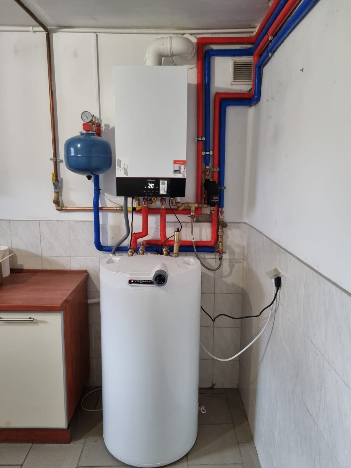 Biały zasobnik ciepłej wody użytkowej Viessmann i wiszący kocioł grzewczy z podłączeniami rur miedzianych i plastikowych w kolorach czerwonym i niebieskim, zamontowane w pomieszczeniu...