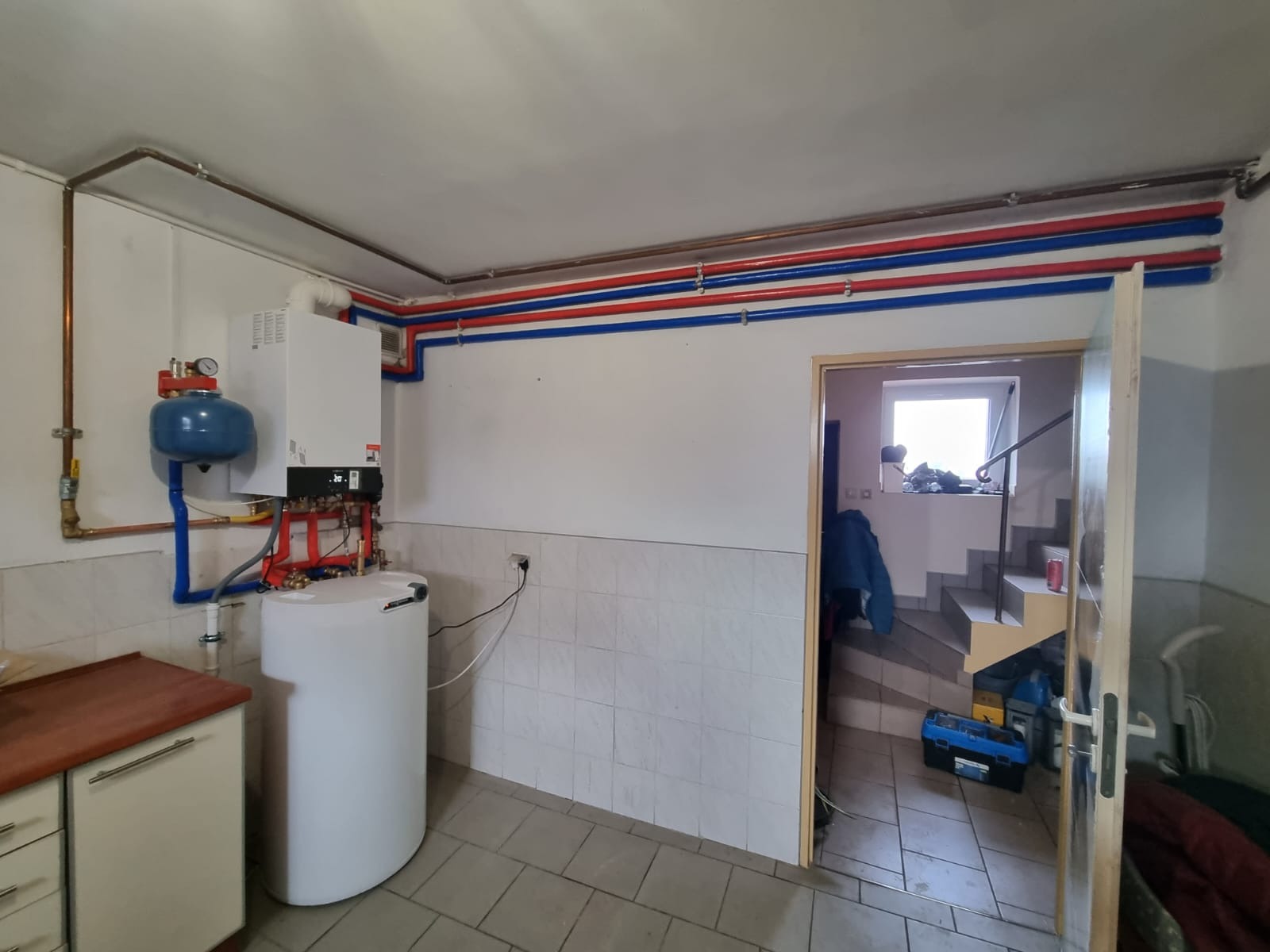 Biały bojler z podłączonymi rurami w kotłowni, widoczne rury miedziane i plastikowe w kolorze czerwonym i niebieskim, w tle schody i skrzynka z narzędziami.