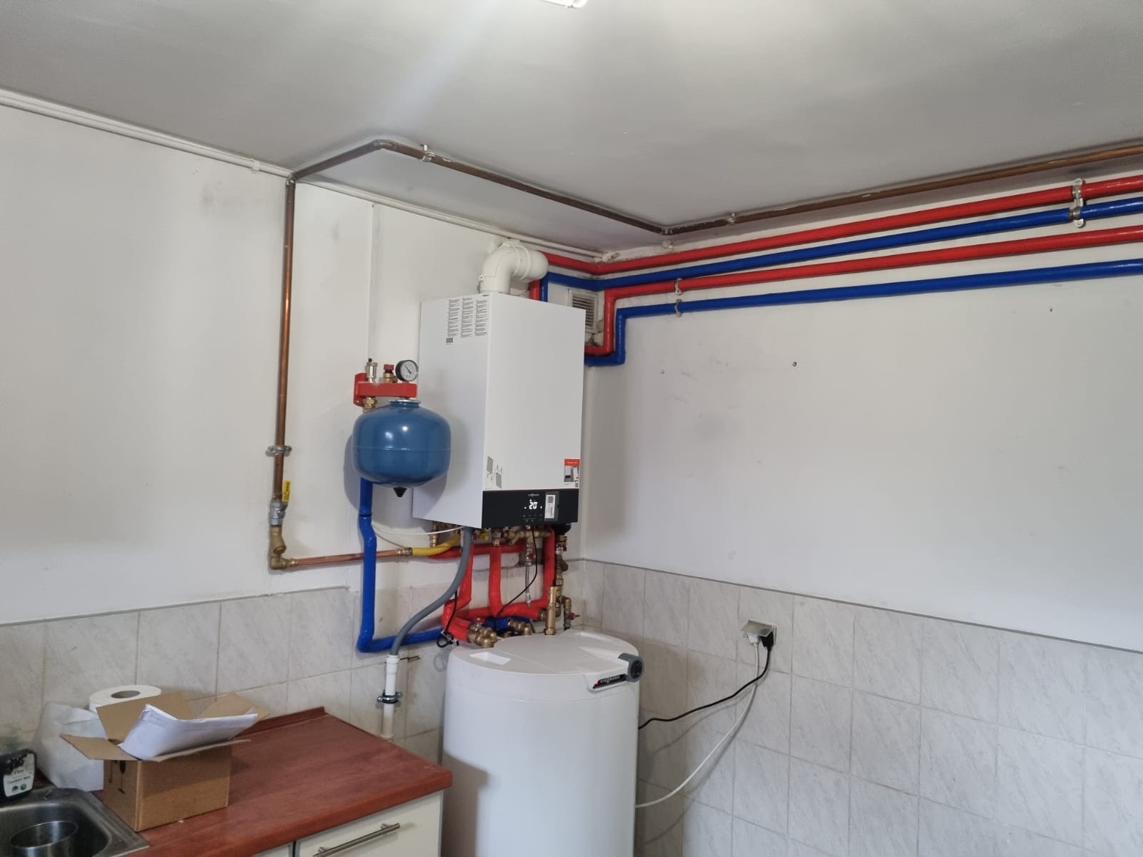 Biały piec centralnego ogrzewania z niebieskim zbiornikiem wyrównawczym i rurami miedzianymi oraz niebiesko-czerwonymi, zamontowany na ścianie wyłożonej płytkami, w pomieszczeniu z niezabudowaną...