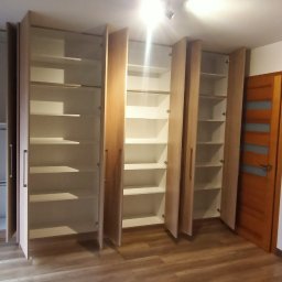 DTM - Wnętrze pokoju z trzema szafami wnękowymi o jasnym, drewnianym wykończeniu, z częściowo otwartymi drzwiami ukazującymi puste półki, na podłodze panele imitujące drewno, w tle widoczne okno...