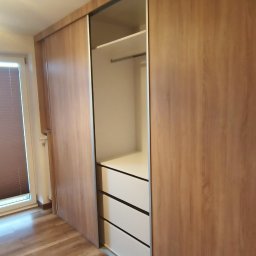 DTM - Wnętrze szafy wnękowej z przesuwnymi drzwiami w kolorze jasnego drewna, widoczne trzy białe szuflady i półka z drążkiem na ubrania.