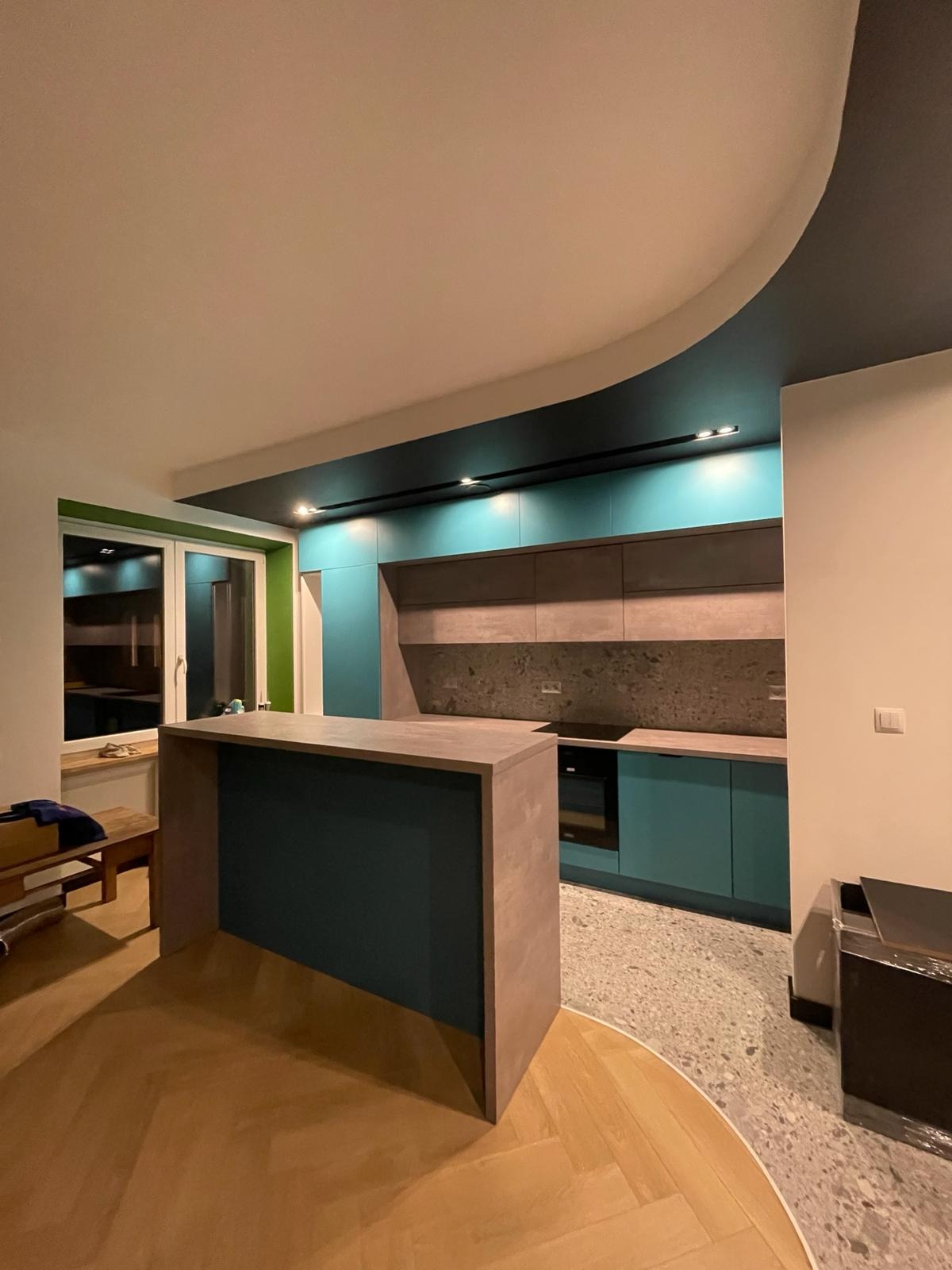 Nowoczesna kuchnia z wyspą kuchenną w kolorze turkusowym i szarym, z drewnianą podłogą w jodełkę i nietypowym sufitem z oświetleniem punktowym.