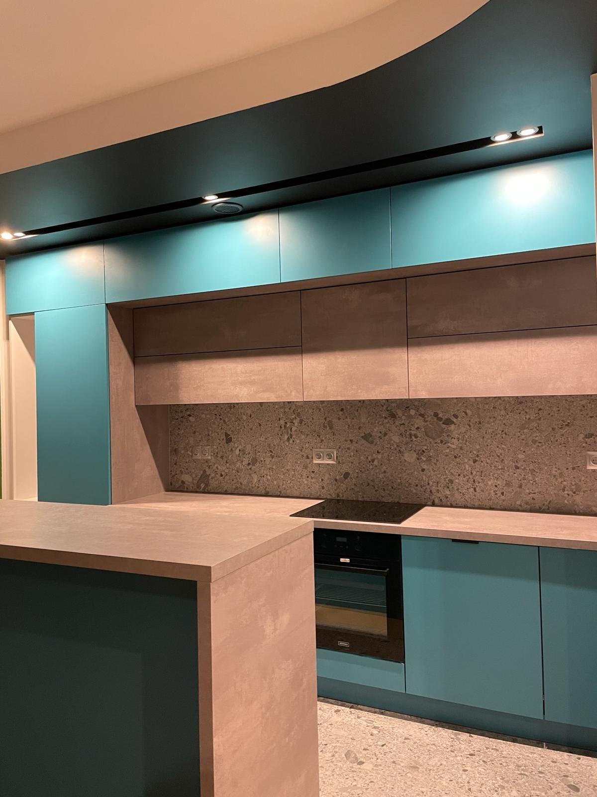 Nowoczesna kuchnia z turkusowymi szafkami górnymi i dolnymi, szarym blatem oraz ciemnoszarym sufitem z oświetleniem punktowym.