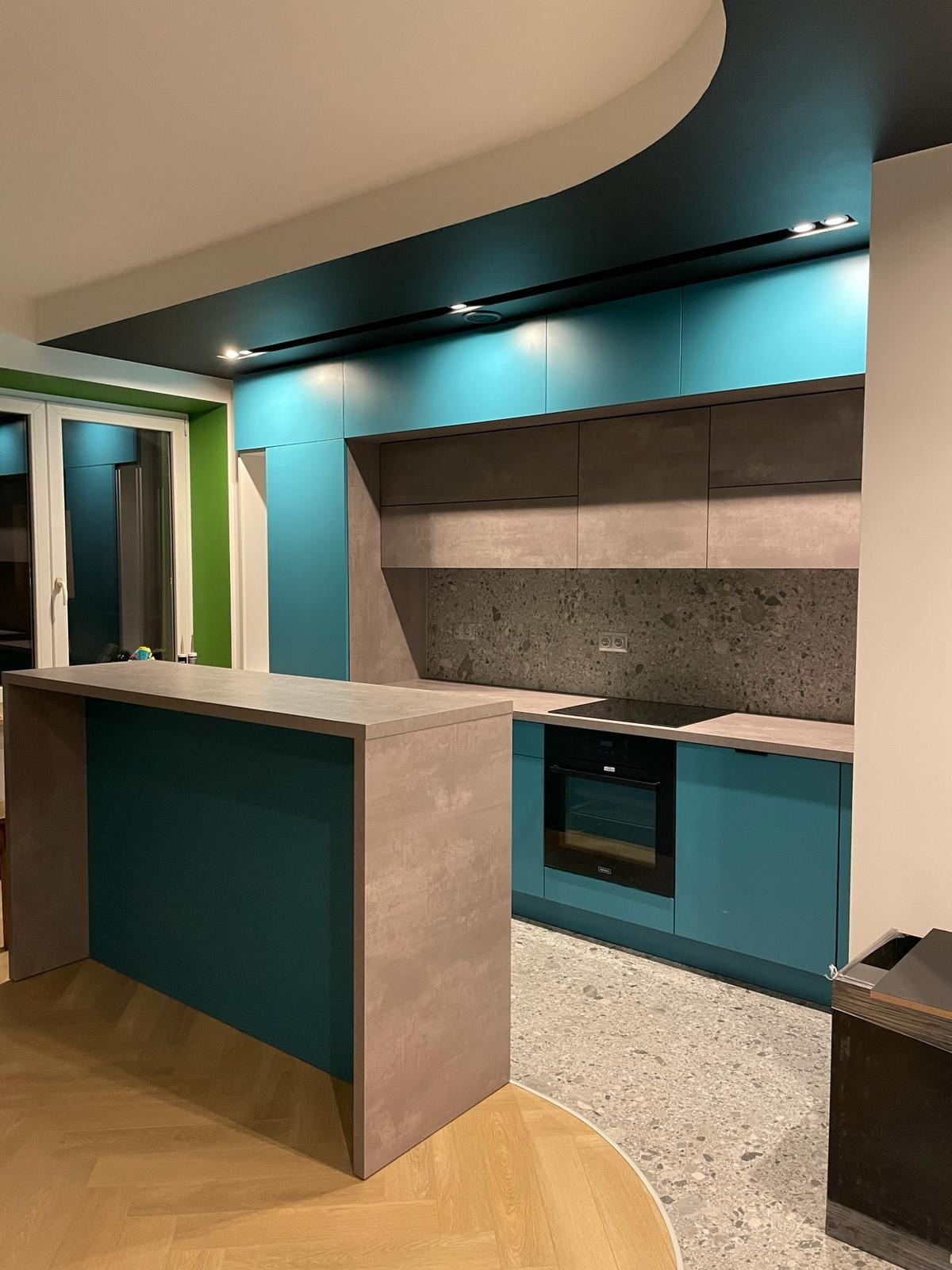 Nowoczesna kuchnia z turkusowymi szafkami, szarym blatem i wyspą, oświetleniem punktowym na czarnym suficie podwieszanym i podłogą z lastryko.