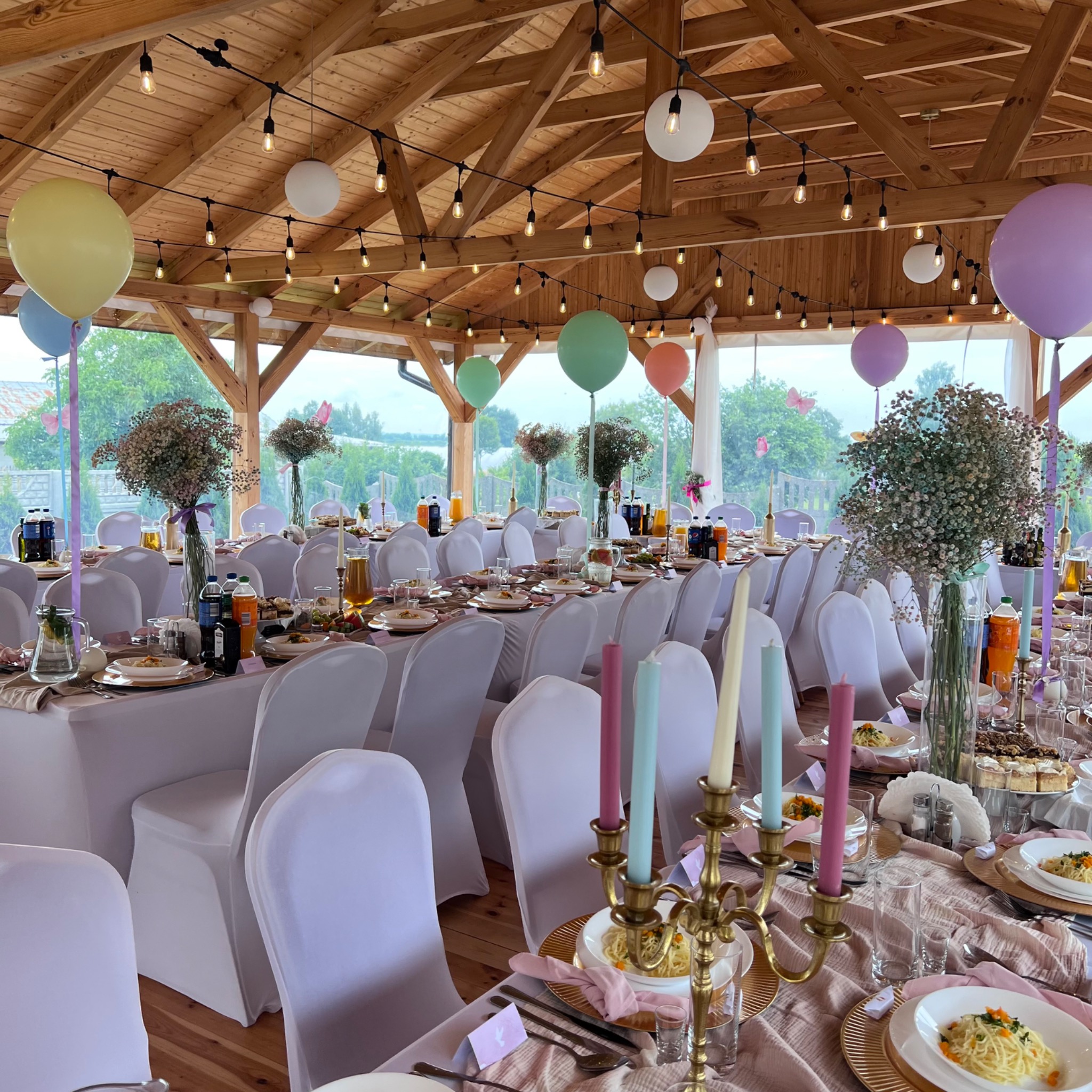 Uroczystość weselna: Długi stół z białymi krzesłami, nakryty różowym obrusem, złote talerze, świeczniki z pastelowymi świecami, balony i kwiaty w szklanych wazonach pod drewnianym zadaszeniem...