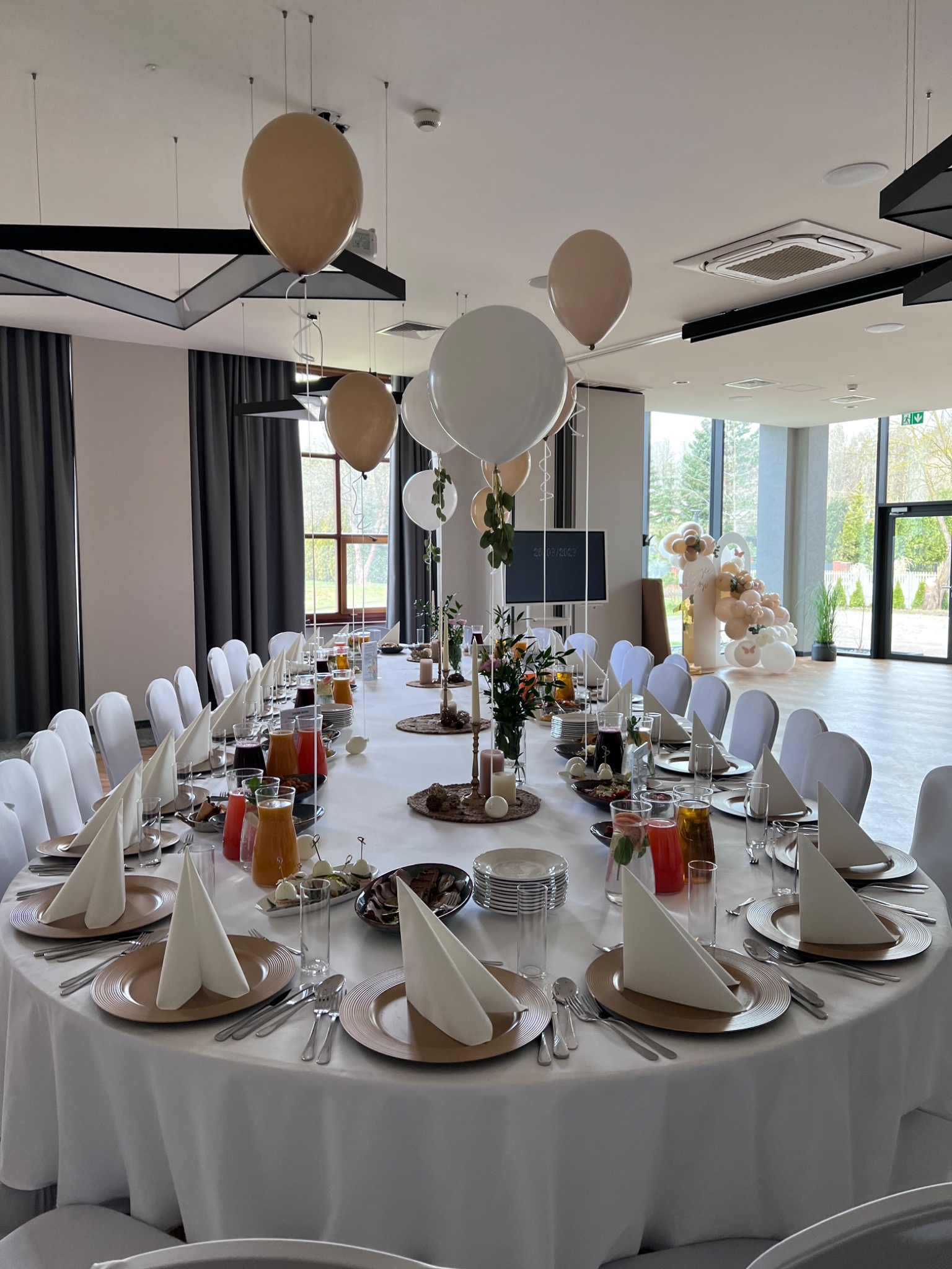 Uroczysty stół weselny z białym obrusem, złotymi talerzami, białymi serwetkami, napojami w szklanych dzbankach i dekoracjami kwiatowymi, nad nim wiszące balony w odcieniach bieli i złota, w tle...