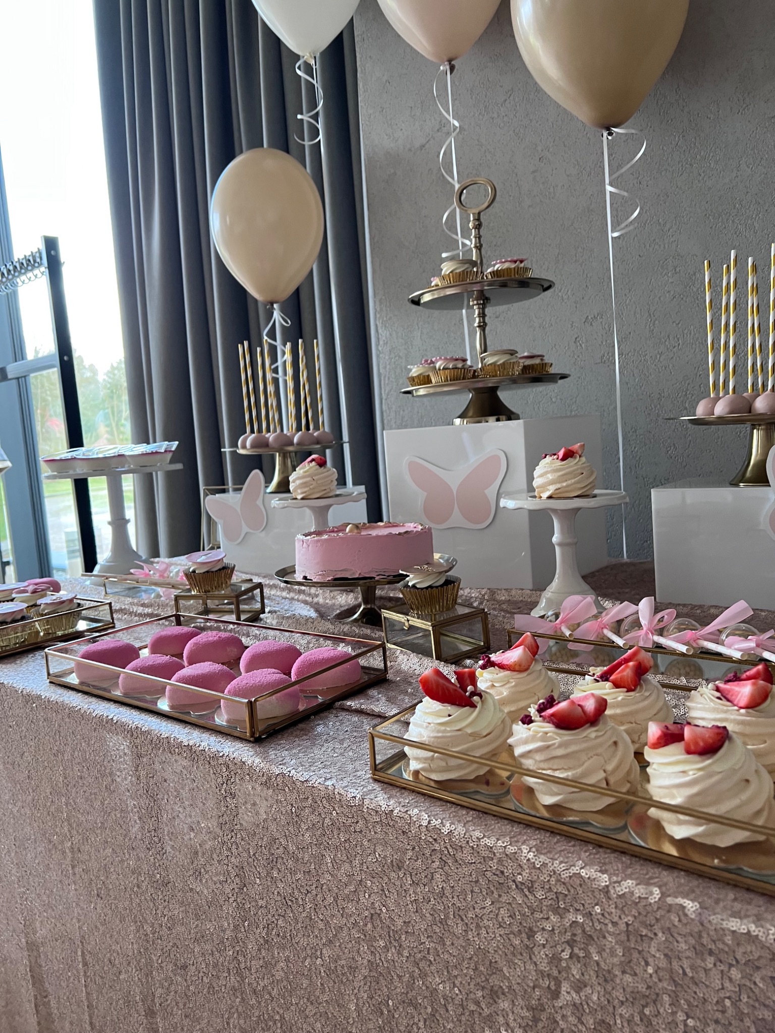 Uroczysty stół z różowymi słodkościami: tort, bezy z truskawkami, babeczki i lizaki, udekorowany balonami i motylami, na błyszczącym obrusie.
