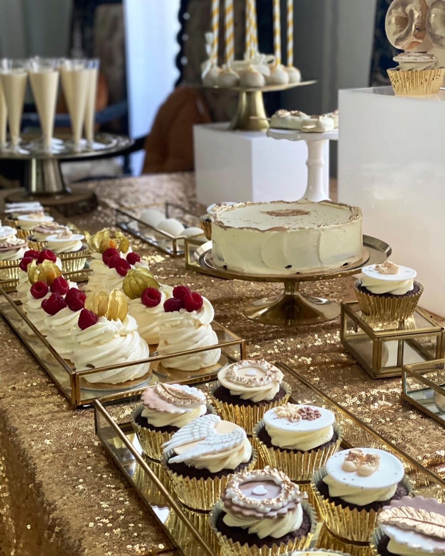 Dekoracyjny stół z ciastami i deserami, w tym babeczki z kremem i ozdobami, bezy z owocami, tort na złotym stojaku, całość na błyszczącym, złotym obrusie.